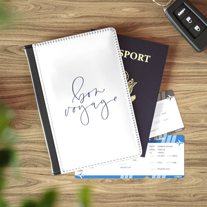 The white front cover of a faux leather passport cover is printed with the phrase 'bon voyage' in a romantic lavender-coloured script and has a black spine.