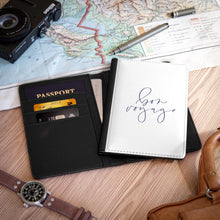 Load image into Gallery viewer, The white front cover of a faux leather passport holder is printed with a lavender coloured script saying 'bon voyage' and has a black spine.