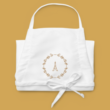 Load image into Gallery viewer, Folded white canvas apron with a graphic of the Eiffel Tower surrounded by a wreath of flowers embroidered in gold thread: L'Abeille Française