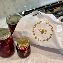Load image into Gallery viewer, A white canvas apron with a graphic of a bumble bee surrounded by a wreath of flowers embroidered in gold thread: L'Abeille Française