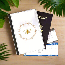 Load image into Gallery viewer, The white front cover of a faux leather RFID passport cover is printed with a peach & gold graphic of a bee surrounded by a wreat of flowers and has a black spine.