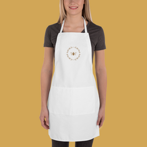 A white canvas apron with a graphic of a bumble bee surrounded by a wreath of flowers embroidered in gold thread: L'Abeille Française
