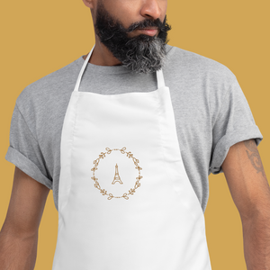 Bib of a white canvas apron with a graphic of the Eiffel Tower surrounded by a wreath of flowers embroidered in gold thread: L'Abeille Française