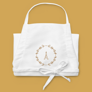 Folded white canvas apron with a graphic of the Eiffel Tower surrounded by a wreath of flowers embroidered in gold thread: L'Abeille Française