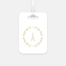 Load image into Gallery viewer, Hard, white plastic luggage tag with a graphic of the Eiffel Tower surround by a wreath of flowers printed on it in gold