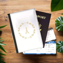 Load image into Gallery viewer, The white front cover of a faux leather passport holder is printed with a gold graphic of the Eiffel Tower surrounded by a wreath of flowers and has a black spine..