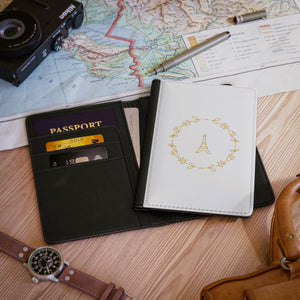 The white front cover of a faux leather passport holder is printed with a gold graphic of the Eiffel Tower surrounded by a wreath of flowers and has a black spine..