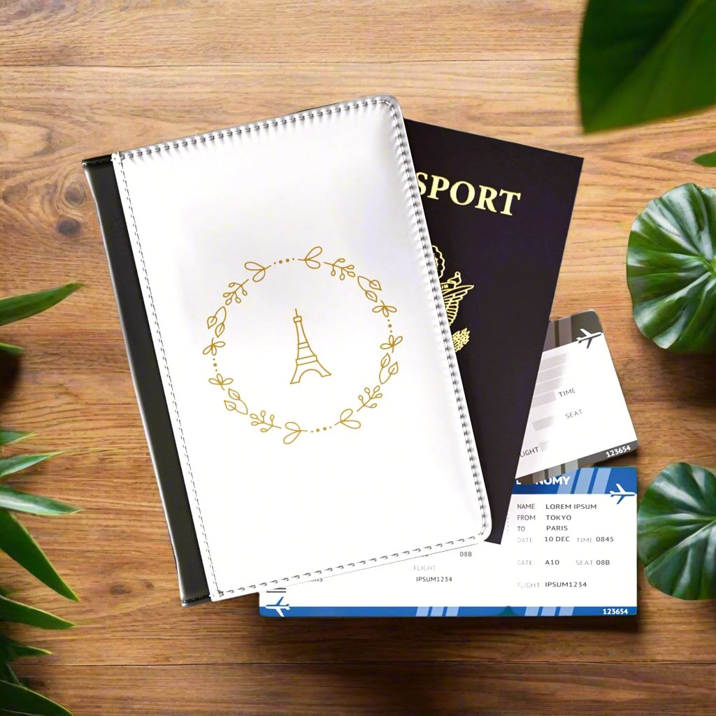 The white front cover of a faux leather passport holder is printed with a gold graphic of the Eiffel Tower surrounded by a wreath of flowers and has a black spine..