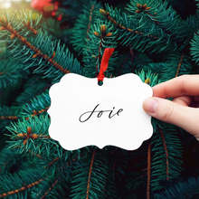 Load image into Gallery viewer, Hand holding a &#39;Joie&#39; ornament against a Christmas tree background