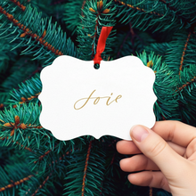 Load image into Gallery viewer, Hand holding a &#39;Joie&#39; ornament against a Christmas tree background