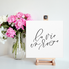 Load image into Gallery viewer, A 10&quot; x 10&quot; square white canvas with the phrase &#39;la vie en rose&#39; printed on it in a contemporary caligraphic script: L&#39;Abeille Française