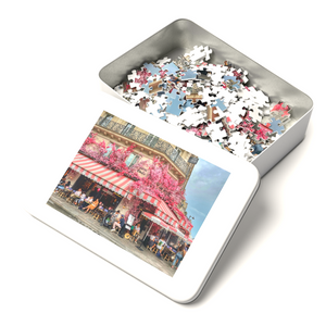 A white tin box containing a 252-piece 14" × 11" jigsaw puzzle of the Restaurant La Favorite in Paris bedecked in pink and white striped awnings and festooned with an abundance of pink flowers. 