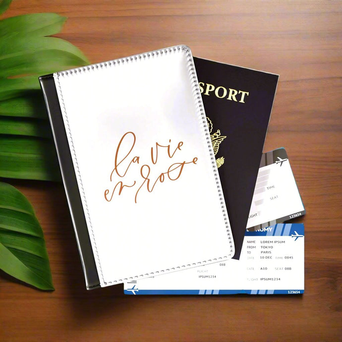 The white front cover of a faux leather passport holder is printed with a rose gold coloured script saying 'la vie en rose' and has a black spine.