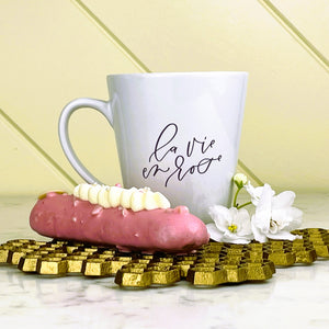 White mug with 'La vie en rose' text on a gold coaster with a pink éclair and white flowers: 'Abeille Française