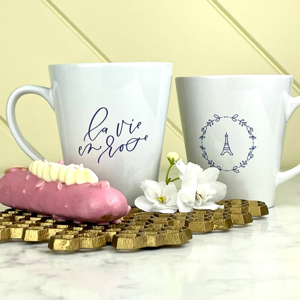 La Vie En Rose Eiffel Latte Mug: èla vie en roseè in lavender script on one side of a white latte mug and a graphic of the Eiffel tower surrounded by a wreath of flowers on the other side: Boutique L'Abeille Française