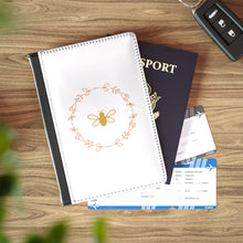 Load image into Gallery viewer, The white front cover of a faux leather RFID passport cover is printed with a peach &amp; gold graphic of a bee surrounded by a wreat of flowers and has a black spine.