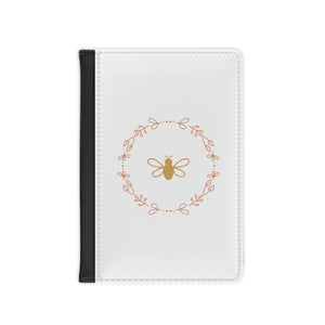 The white front cover of a faux leather RFID passport cover is printed with a peach & gold graphic of a bee surrounded by a wreat of flowers and has a black spine.