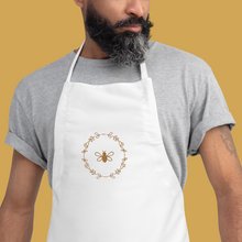 Load image into Gallery viewer, A white canvas apron with a graphic of a bumble bee surrounded by a wreath of flowers embroidered in gold thread: L&#39;Abeille Française