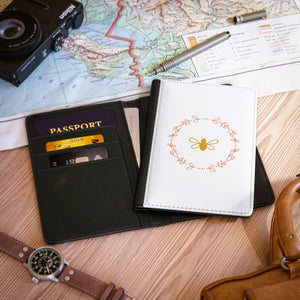 The white front cover of a faux leather RFID passport cover is printed with a peach & gold graphic of a bee surrounded by a wreat of flowers and has a black spine.