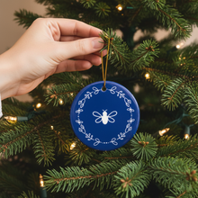 Load image into Gallery viewer, Hand holding a blue ornament with a white bee design on a Christmas tree.