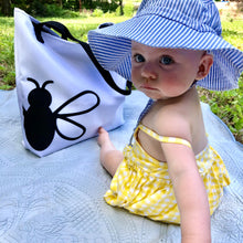 Load image into Gallery viewer, An infant in a sunhat sits on a blanket beside the Wandering Bee Tote. This tote has a white front with 3 quarters of a black stylized bee printed on lower left side, black stitching and webbing handles: Boutique L&#39;Abeille Française