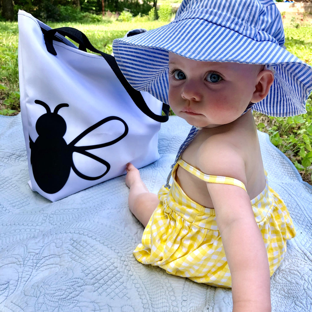 An infant in a sunhat sits on a blanket beside the Wandering Bee Tote. This tote has a white front with 3 quarters of a black stylized bee printed on lower left side, black stitching and webbing handles: Boutique L'Abeille Française