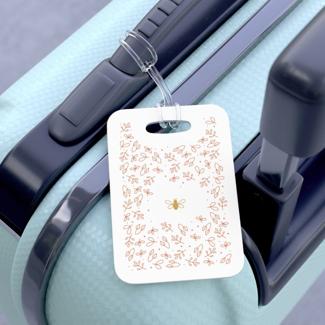 Hard, white plastic luggage tag with gold bee surrounded by pink flowers printed on it 