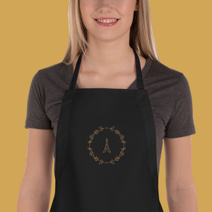 Bib of a black canvas apron with a graphic of the Eiffel Tower surrounded by a wreath of flowers embroidered in gold thread: L'Abeille Française