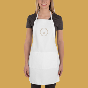 A white canvas apron with a graphic of the Eiffel Tower surrounded by a wreath of flowers embroidered in gold thread: L'Abeille Française