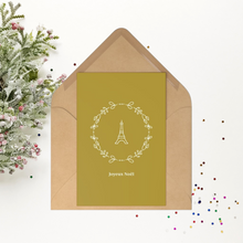 Load image into Gallery viewer, Gold rectangular Christmas postcard with a wreathed Eiffel Tower graphic and the phrase Joyeux Noel printed in off-white set against an open kraft paper envelope.