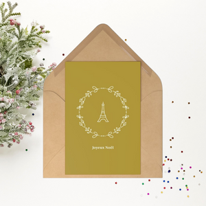 Gold rectangular Christmas postcard with a wreathed Eiffel Tower graphic and the phrase Joyeux Noel printed in off-white set against an open kraft paper envelope.