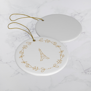 Glossy, round, white ceramic ornament decorated with a gold-toned Eiffel Tower and wreath motif lying next to the white back of another