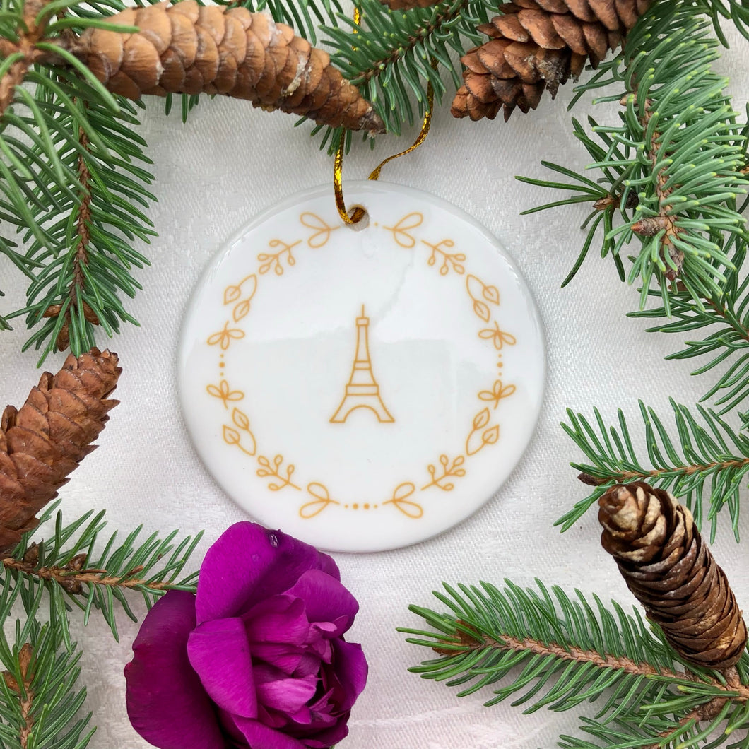 Glossy, round, white ceramic ornament decorated with a gold-toned Eiffel Tower and wreath motif