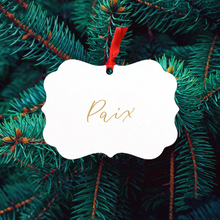 Load image into Gallery viewer, A glossy white ornament with the word &#39;Paix&#39; printed in gold, hanging from a red satin ribbon on a green pine tree.