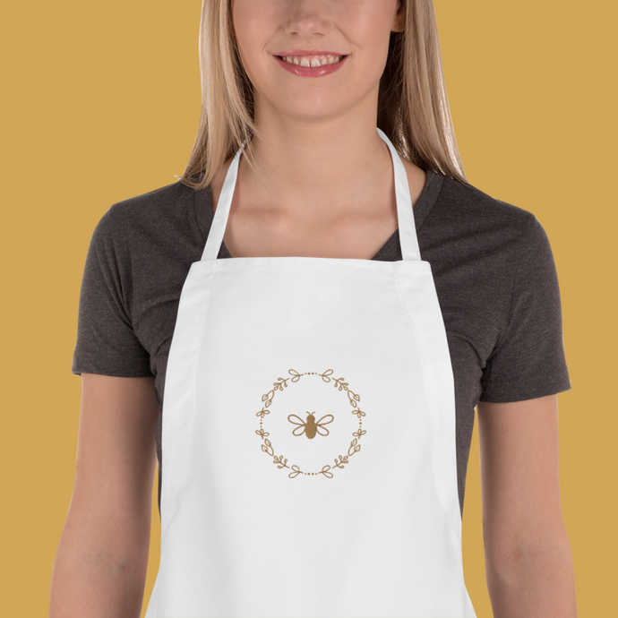 Bib of a white canvas apron with a graphic of a bumble bee surrounded by a wreath of flowers embroidered in gold thread: L'Abeille Française
