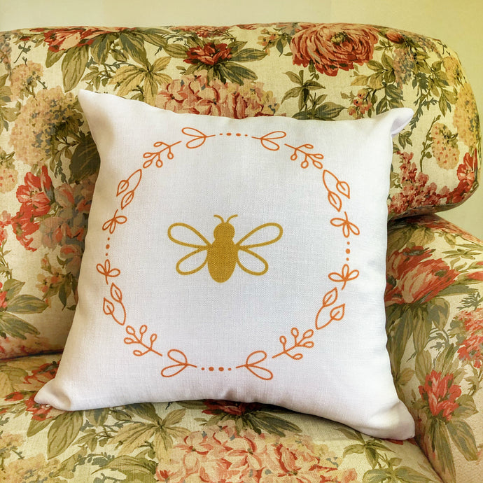 A white cushion with a gold bee surrounded by a rose gold wreath of flowers graphic sitting on a warm floral print chesterfield
