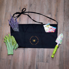 Load image into Gallery viewer, Black garden apron with three pockets, two ties and a gold bee and wreath graphic