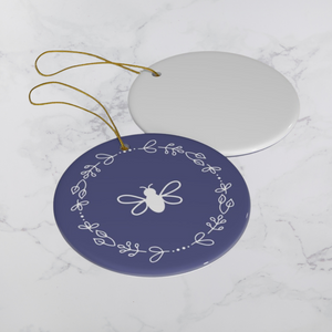 Glossy, round, lavender-coloured ceramic ornament with a white bee and wreath motif next to the white back of another